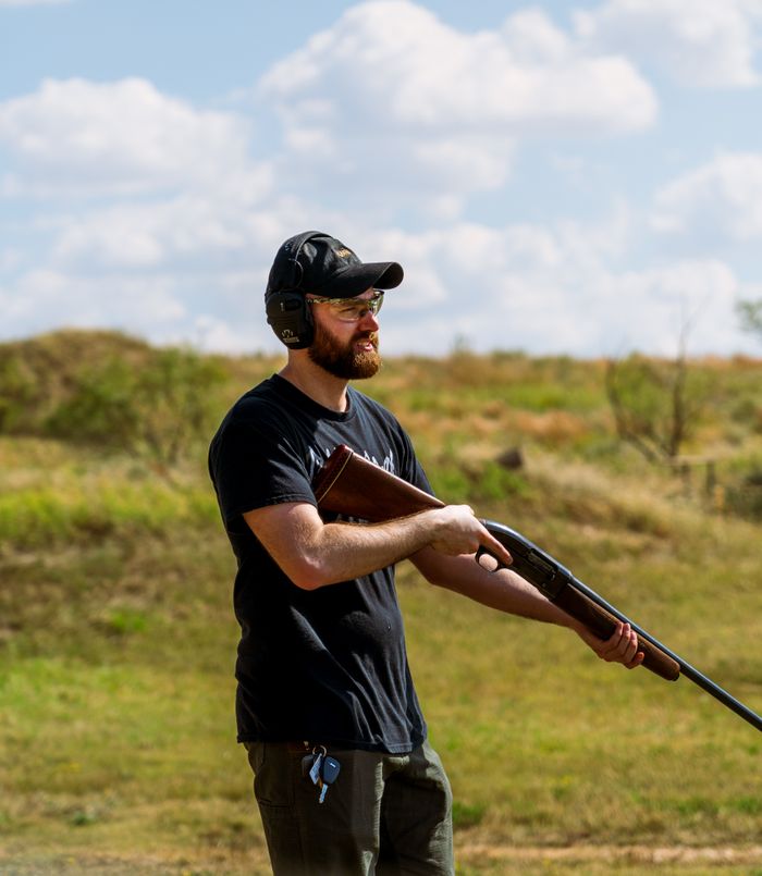 Bearded man in protective gear holding a shotgun in an outdoor field
