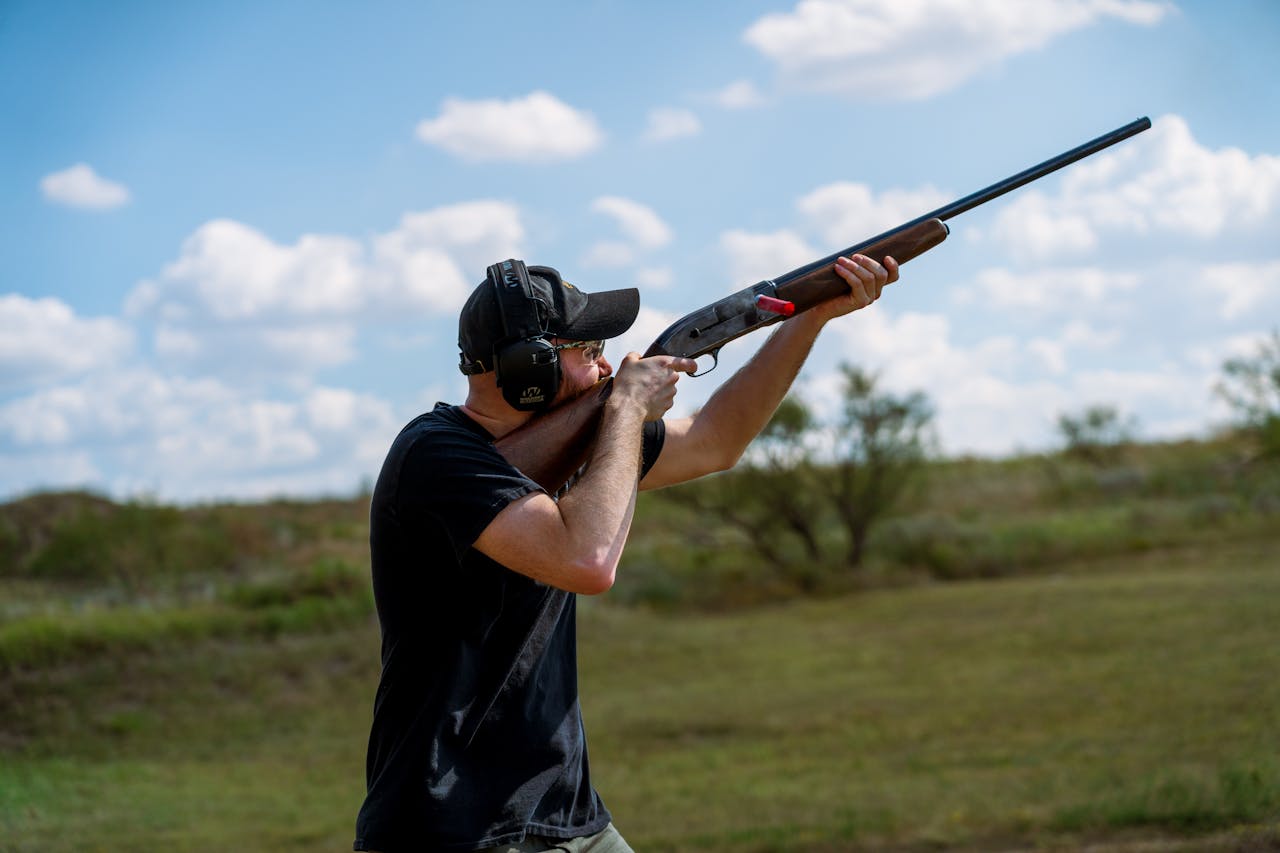 A man aiming a rifle outdoors with ear protection against a blue sky with clouds.