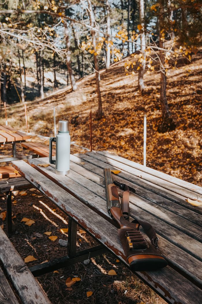 Wooden picnic table in autumn forest, featuring a rifle and thermos, conveying an outdoor adventure vibe.
