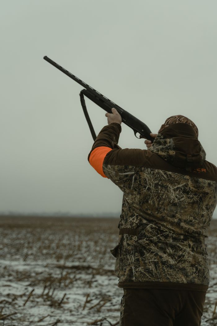 Hunter in camouflaged gear aiming a shotgun in winter field.