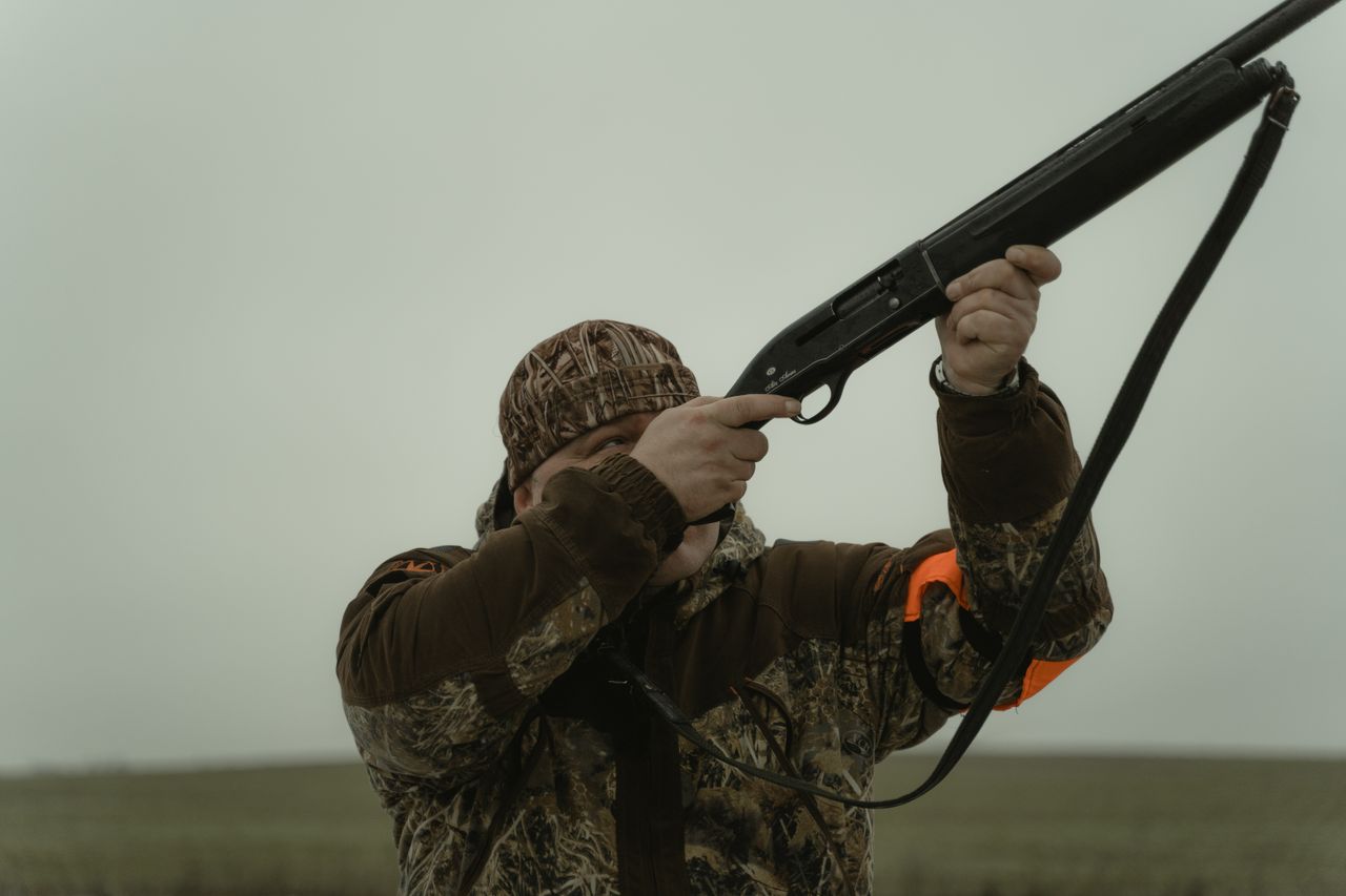 Hunter wearing camouflage gear aiming a shotgun in an open field setting, demonstrating outdoor hunting activity.
