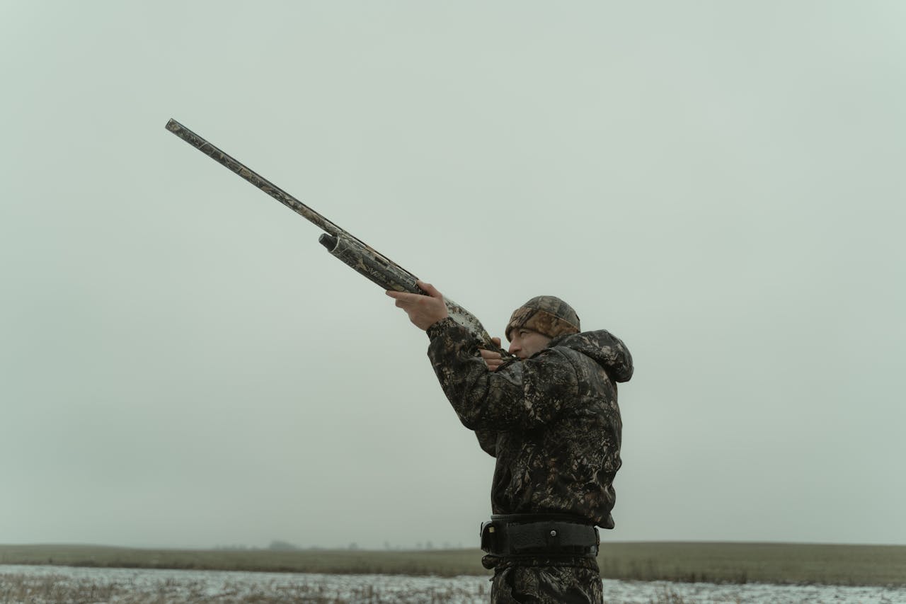 A hunter in camouflage aiming a shotgun in an open field, capturing the essence of outdoor sport.